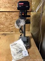 Full view of Craftsman 8-inch drill press including manual and mounting hardware on the base, showing drill press upright with power switch visible.