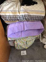 Photo showing stacked bedding including comforters with geometric and floral patterns and a light purple sheet on top.