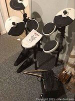 Full kit with five drum pads, two cymbal pads, metal rack, foot pedals on floor, drumsticks laid on pedals