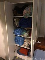 Tall white cabinet door open revealing shelves with assorted folded linens, sleeping bags, and towels.