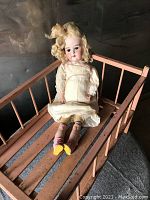 Full view of German doll seated on the wooden toy bed showing dress and shoes, and bed with slatted sides.