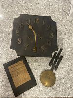 Top view of the wooden clock case, clock face with brass numerals and hands, a detached pendulum, and a wooden panel with label.