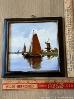 Front view of the hand painted Iris Gouda tile framed, showing a sailing scene with boats and a windmill, with a ruler for scale measuring approximately 7 x 7 inches