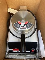 Closed waffle maker in original box showing stainless steel housing, control panel and power cord coiled on top