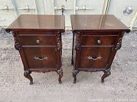 Front view of both wooden bedside tables side by side showing carved legs, ornamental appliqués, cabinet doors with hardware