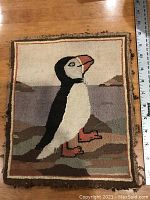Front view of Grenfell hooked mat depicting a puffin standing on rocky terrain with ruler showing size about 10 by 12 inches.