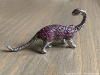 Front side view of the silver-tone dinosaur brooch with red rhinestones on the body and eyes, showing detailed texture and shape.