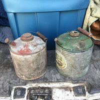 Pair of vintage metal kerosene cans with wooden handles, placed side by side on a vehicle floor with blue storage bin in the background.