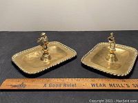 Photo showing two rectangular brass ashtrays side by side with ruler for scale. Both have a twisted rope border and a figurine mounted in the center; one playing flute, one standing man figurine.