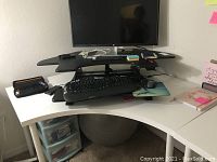 White corner desk table viewed from side with black multi-level monitor stand and computer accessories on top.