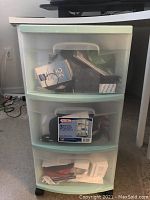Medium sized three drawer plastic storage cart with light green trim, translucent drawers with various items inside under a table.
