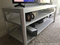 White Ikea TV unit with a metal frame and three shelves, holding various multimedia and decor items, placed on carpeted floor near window.