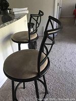Side view of two black metal bar stools with padded beige seats and curved armrests, placed next to a granite countertop.