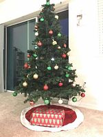 Full view of the decorated 6.5 foot artificial Christmas tree with multicolor lights and round ornaments, tree skirt, and gift box beneath
