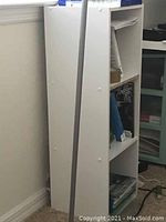 Side view of a white bookshelf featuring three shelves, positioned upright beside a wall and carpet flooring.