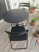 Table and two chairs set shown on wooden balcony with potted plants around.