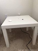 White small square IKEA side table with flat top and straight legs set against beige carpet and white wall with electrical outlets.