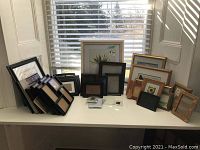 Wide angle photo of multiple picture frames arranged on a white table by a window. Includes black and wood-finished frames with magnetic holders in front.