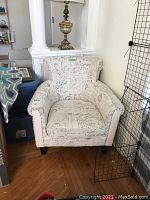 Front angled view of French themed upholstered armchair showing the script patterned fabric, general shape, and caster wheels.