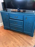 Front view of blue painted TV cabinet with three drawers and two carved door panels, original brass hardware visible.