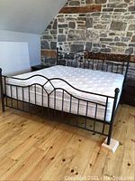 Black metal hollow tube bedframe fully assembled with mattress, set against a stone wall and wooden floor.