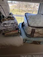 View of stacked bedding including folded comforter, coverlets, patterned pillow, and decorative pillow on a bed by a window