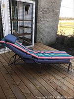 Side view of chaise lounge on wooden deck showing striped cushion and metal frame with plastic webbing and adjustable back.