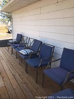 Six metal patio chairs with blue cushions lined up on wooden deck against siding wall and red door.