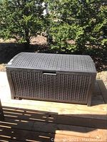 Full view of brown resin woven patio storage box on wooden deck, curved lid, closed position.