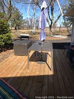 Photo showing metal slatted patio table with mounted blue weathered umbrella on a wooden deck, outdoor setting.