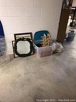 Wide shot showing storage container with picture frames, small wooden wine rack on top, assorted framed prints leaning against the wall