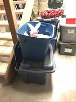 Stacked storage bins including a large blue plastic bin and smaller black bin, photographed indoors near stairs