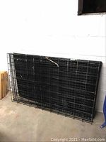 Folded black metal dog crate with floor panel leaning against a wall on a concrete floor.