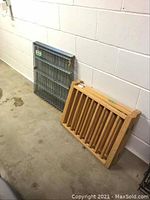 Two collapsed pet pens side by side against a white wall on concrete floor: one wooden and one metal.