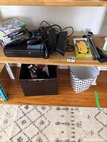 Front shelf view showing Xbox 360 console, one wired controller, Kinect sensor, power adapter, six game cases, partial view of phone case packaging and tripod legs.