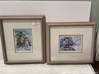 Two framed prints by Nori Peters, side by side on a table, showing Inuit/northern scenes with people and dogs in snow.