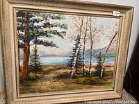 Framed oil painting showing a landscape of trees, water, and mountains with visible frame damage and scratches.