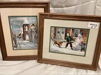 Two framed prints displayed leaning against a surface, showing one print with children and a snowman, the other with a snow-covered street scene.