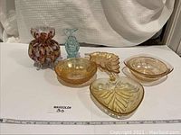 Overview of 6 assorted glassware pieces arranged on white surface with fabric background. Includes various amber/golden colored bowls and dishes, a multicolored ruffled vase, and a blue glass fish figurine.