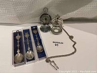 All items displayed together: pocket watch with chain, open compass, and three collector spoons in individual boxes