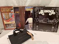 Five music album vinyl records arranged standing behind a Mary Kay makeup bag on a flat surface, with a gold-tone wristwatch placed on the makeup bag.