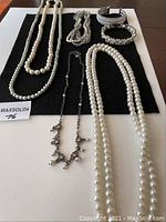 Seven pieces of costume jewelry laid out on a black cloth including five necklaces and two bracelets, mainly featuring faux pearls and rhinestones.