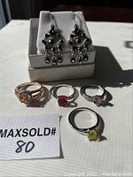 Four rings displayed on a surface with an auction lot number card, showing different colored stones: amber, red, pink heart-shaped, and yellow; rings approximate size 8.