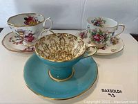 Three vintage tea cups with matching saucers displayed together. The turquoise Aynsley cup is centered in front, flanked by two floral patterned cups with scalloped saucers behind.