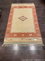 Full view of the rectangular flatwoven throw rug laying flat on dark hardwood floor showing terracotta border and central diamond medallion on cream field.