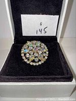 Round brooch displayed upright in a box, showing top view with rhinestones and gold-tone metal backing.