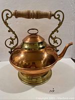 Full view of the ornate copper coloured kettle with a wooden handle and curved spout.