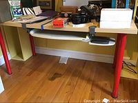 Full view of the desk with assorted items on top, showing the keyboard tray underneath and red metal legs touching the hardwood floor. Scratches and wear visible on the surface.