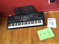 Front view of black Casio CTK-541 keyboard on wood floor with attached clear music stand and manuals beside it.