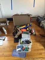Overview photo showing multiple plastic bins and boxes containing various toys including Nerf blaster, model airplanes, and assorted small figures.